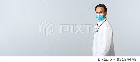 Covid-19, virus, healthcare workers and vaccination concept. Profile of professional male doctor in medical mask turn head at camera and look confident. Promo of clinic services, medication 85184448