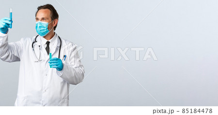 Covid-19, preventing virus, healthcare workers and vaccination concept. Serious professional med lab doctor in medical mask and gloves looking at test-tube with coronavirus vaccine 85184478