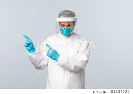 Covid-19, preventing virus, healthcare workers and vaccination concept. Serious doctor in personal protective equipment pointing fingers upper left corner at information banner Covid-19, preventing virus, healthcare workers and vaccination concept. Serious doctor in personal protective equipment pointing fingers upper left corner at information banner 85184520