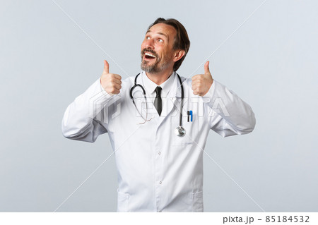 Covid-19, coronavirus outbreak, healthcare workers and pandemic concept. Enthusiastic handsome doctor in white coat looking at top left advertisement and thumbs-up, recommend product 85184532