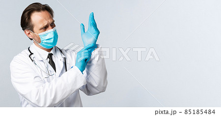 Covid-19, preventing virus, healthcare workers and vaccination concept. Serious professional doctor in medical mask put on gloves before examining patient in clinic, working in hospital 85185484