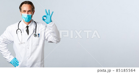 Covid-19, preventing virus, healthcare workers and vaccination concept. Clinic doctor in medical mask and gloves provide best quality service, smiling and showing okay gesture Covid-19, preventing virus, healthcare workers and vaccination concept. Clinic doctor in medical mask and gloves provide best quality service, smiling and showing okay gesture 85185564