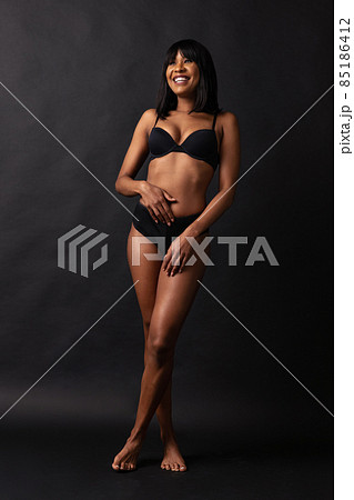 Full-length portrait of young beautiful slim girl in black underwear standing isolated over dark studio background. Natural beauty concept. 85186412