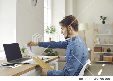 Young man who's sitting at desk in his home office opens envelope with business letter. Serious businessman sitting at table with laptop computer takes and reads sales report that he got by paper mail 85190306