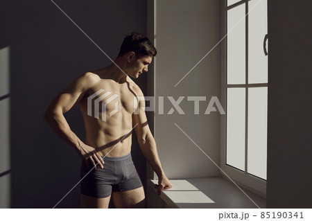 Handsome fit man posing in underwear. Young male model wearing comfortable black cotton underpants standing near window. Studio shot with contrasting bright sunlight and shadows. Male fashion concept Handsome fit man posing in underwear. Young male model wearing comfortable black cotton underpants standing near window. Studio shot with contrasting bright sunlight and shadows. Male fashion concept 85190341