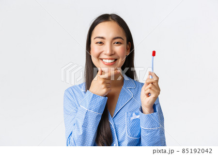 Close-up of attractive asian woman in blue pajama thinking while brushing her teeth with toothbrush, smiling and touching chin pondering great idea, white background Close-up of attractive asian woman in blue pajama thinking while brushing her teeth with toothbrush, smiling and touching chin pondering great idea, white background 85192042
