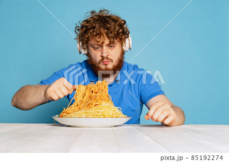 Emotional young hairy red-bearded man tasting large portion of noodles, pasta isolated on blue studio background. 85192274
