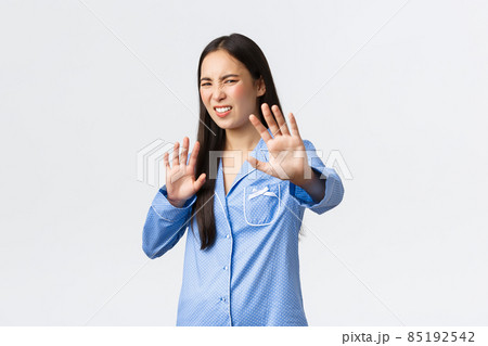 Digusted and displeased asian girl in blue pajamas extend hands forward in refusal, rejecting something disgusting, grimacing from aversion and dislike, shaking hands in denial over white background 85192542