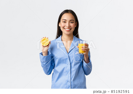 Morning, active and healthy lifestyle and home concept. Smiling cheerful asian girl starting her day with fresh made orange guice, holding glass and half of orange, looking happy and energized Morning, active and healthy lifestyle and home concept. Smiling cheerful asian girl starting her day with fresh made orange guice, holding glass and half of orange, looking happy and energized 85192591