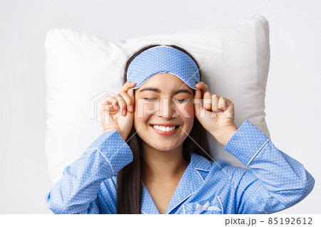Close-up of happy beautiful asian girl in sleeping mask and blue pajamas lying in bed, smiling upbeat, having perfect mood as going bed and getting ready sleep, standing white background Close-up of happy beautiful asian girl in sleeping mask and blue pajamas lying in bed, smiling upbeat, having perfect mood as going bed and getting ready sleep, standing white background 85192612