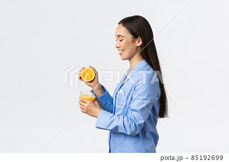 Morning, active and healthy lifestyle and home concept. Profile or beautiful healthy asian girl in blue pajamas squeezing orange juice in glass and smiling happy, starting day right 85192699