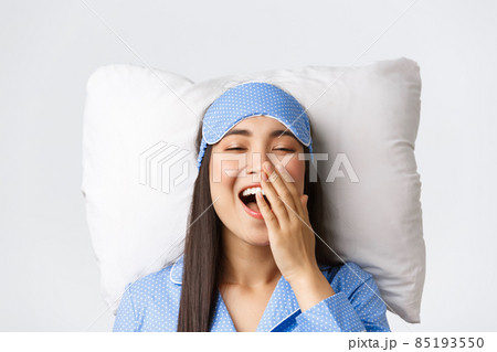 Close-up of happy smiling asian girl lying in bed on pillow wearing blue pajamas and sleeping eyemask, yawning satisfied after good night sleep, posing over white background 85193550