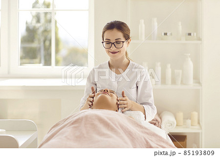 Portrait of female cosmetologist doctor doing massage and beauty treatments in beauty salon. Smiling beautician massages face of female client lying on massage table and covered with towel. Portrait of female cosmetologist doctor doing massage and beauty treatments in beauty salon. Smiling beautician massages face of female client lying on massage table and covered with towel. 85193809