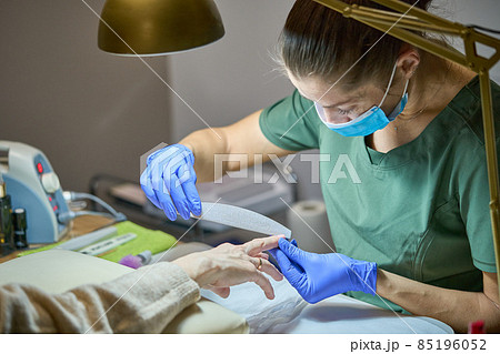 Manicurist filing client nail with a nail file. Manicure process in beauty salon. Master in blue gloves, mask and green uniform under the gold lamp. Manicurist filing client nail with a nail file. Manicure process in beauty salon. Master in blue gloves, mask and green uniform under the gold lamp. 85196052