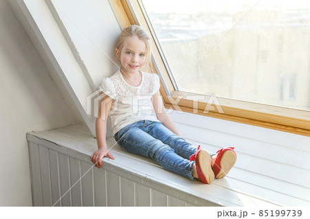 Little cute sweet smiling girl in jeans and white T-shirt sitting on the window sill in bright light living room at home and thinking Childhood, schoolchildren, youth, relax concept Little cute sweet smiling girl in jeans and white T-shirt sitting on the window sill in bright light living room at home and thinking Childhood, schoolchildren, youth, relax concept 85199739
