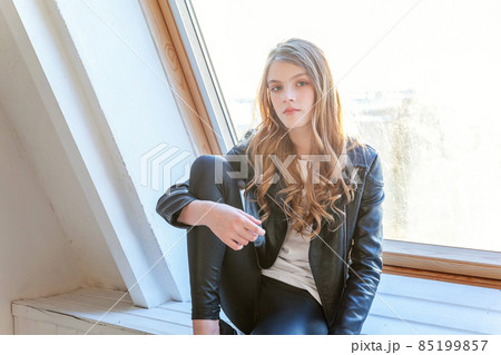 Young teenage girl in leather jacket sitting on modern chair posing in bright light room against white wall background window. Pretty sexy fashion sensual woman dressed in hipster rock style outfit Young teenage girl in leather jacket sitting on modern chair posing in bright light room against white wall background window. Pretty sexy fashion sensual woman dressed in hipster rock style outfit 85199857