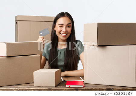 Small business owners, startup and e-commerce concept. Successful asian businesswoman showing credit card, except contactless payment while selling products online, wink cheeky and smiling 85201537