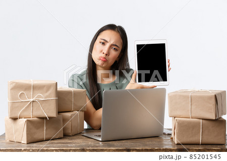 Small business owners, startup and e-commerce concept. Disappointed asian woman pouting upset, showing bad stats on digital tablet screen, using laptop, packing orders in boxes for shipping 85201545