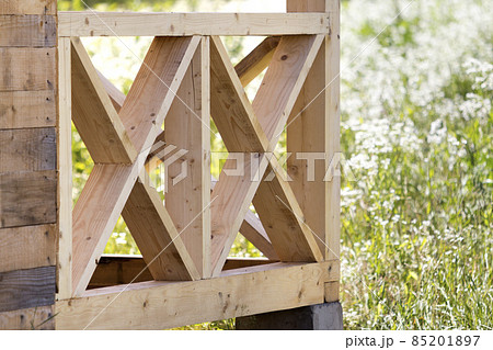 Close-up section geometrical decorative simple detail of new natural not painted wooden planks and bars for balcony, attic fence on blurred blooming summer field background. Construction and design. 85201897