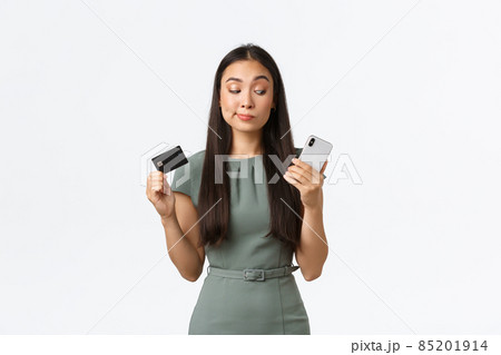 Small business owners, women entrepreneurs concept. Hesitant skeptical asian woman trying new banking application and looking doubtful at smartphone screen while showing credit card Small business owners, women entrepreneurs concept. Hesitant skeptical asian woman trying new banking application and looking doubtful at smartphone screen while showing credit card 85201914