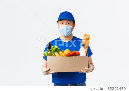 Online shopping, food delivery and covid-19 pandemic concept. Young asian male courier in blue uniform, medical mask and gloves bring grocery order to client home, white background Online shopping, food delivery and covid-19 pandemic concept. Young asian male courier in blue uniform, medical mask and gloves bring grocery order to client home, white background 85202429