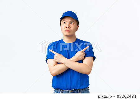 Thoughtful indecisive asian courier in blue uniform thinking, looking unsure while pointing sideways, trying make right choice, standing clueless over white background. Delivery guy got confused Thoughtful indecisive asian courier in blue uniform thinking, looking unsure while pointing sideways, trying make right choice, standing clueless over white background. Delivery guy got confused 85202431