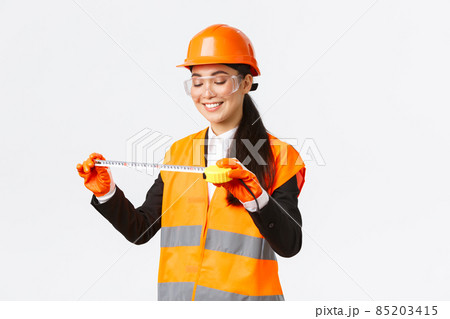 Pleased smiling asian female architect achieve good results, looking satisfied at tape measure after measuring layout at construction area, standing in safety helmet and reflective clothing 85203415