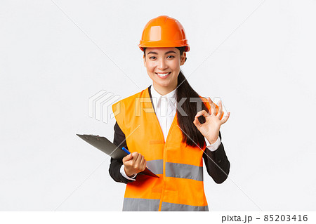 Satisfied smiling asian female architect, construction engineer in helmet and reflective jacket, showing okay gesture, approve and give permission for builing works, finish inspection 85203416