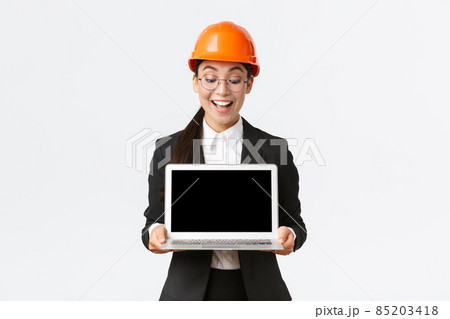 Impressed happy asian female chief engineer showing great results of enterprise to investors, standing in safety helmet and business suit, holding laptop and look pleased at computer screen 85203418