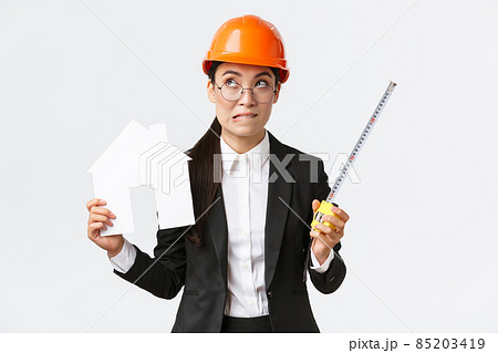 Indecisive asian female architect, construction engineer in helmet and business suit holding house maket and tape measure, biting lip unsure, looking away thoughtful, searching solution 85203419
