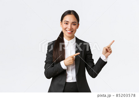 Confident asian female entrepreneur, saleswoman in suit introduce product, showing place to clients. Businesswoman smiling and pointing fingers right at banner, standing white background 85203578