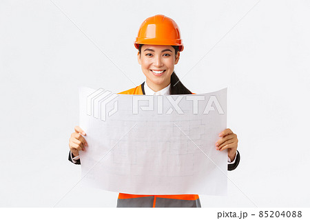 Excited smiling asian female engineer at construction area study blueprints, looking pleased while reading documents, building plan, standing in safety helmet over white background 85204088