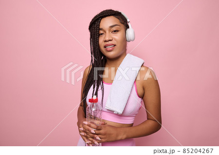 Pleasant young African fitness woman with wireless headphones, dressed in pink tracksuit holding a bottle of water and looking at the camera, isolated on pink background. Sport and recreation concept 85204267