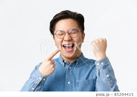 Orthodontics and stomatology concept. Close-up of satisfied happy asian man pointing at his dental braces and smiling broadly, fist pump, rejoicing, fixing teeth, standing white background Orthodontics and stomatology concept. Close-up of satisfied happy asian man pointing at his dental braces and smiling broadly, fist pump, rejoicing, fixing teeth, standing white background 85204415