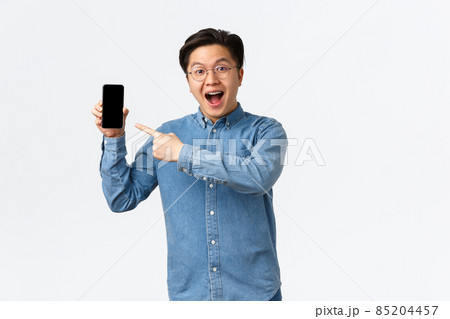 Excited and impressed asian guy in glasses and braces, showing awesome thing on mobile phone screen, pointing finger at smartphone display as introduce new application, white background 85204457