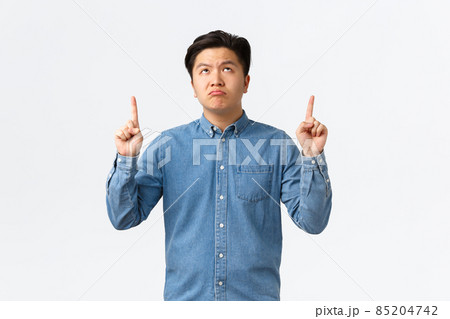 Skeptical and unamused asian man in blue shirt looking and pointing fingers up with disappointed expression, pouting distressed, complaining over something bad, standing white background 85204742