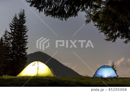 Two brightly lit tourist tents on green grassy forest clearing on dark mountain and clear blue starry sky copy space background. Tourism, night camping in summer mountains, beauty of nature concept. 85205008
