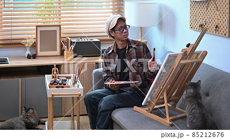 Asian male artist painting picture on easel in living room. Asian male artist painting picture on easel in living room. 85212676