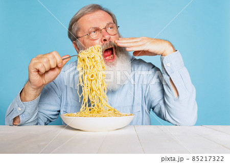 Senior hairy gray-bearded man tasting large portion of noodles, pasta isolated on blue studio background. Senior hairy gray-bearded man tasting large portion of noodles, pasta isolated on blue studio background. 85217322