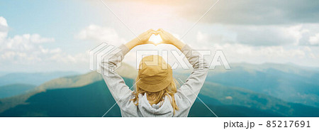 Back view of young traveler female in hat standing over summer mountain peaks and making shape of love heart by hands. Back view of young traveler female in hat standing over summer mountain peaks and making shape of love heart by hands. 85217691