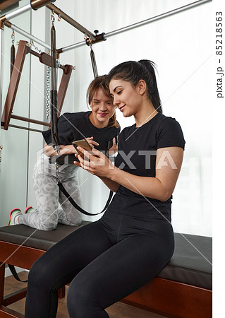 Brunette caucasian woman personal trainer during a workout session on pilates reformer bed equipment with an attractive client in light medical office. Healthy lifestyle rehabilitation concept. 85218563