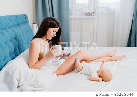 mom sits on the bed with a phone and a cup next to the baby. 85225044