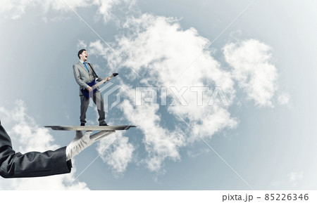 Businessman on metal tray playing electric guitar against blue sky background 85226346
