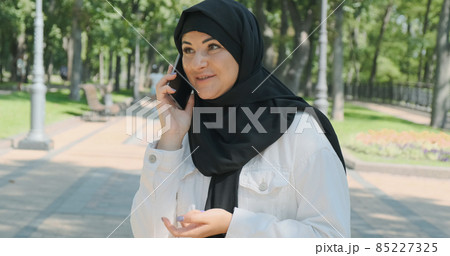Happy arabian woman talking on smartphone on summer city park, close-up. Young female in black headscarf communicates by cellphone in sunny street outdoor. Smiling muslim girl speaking on mobile phone Happy arabian woman talking on smartphone on summer city park, close-up. Young female in black headscarf communicates by cellphone in sunny street outdoor. Smiling muslim girl speaking on mobile phone 85227325