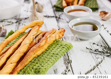 Crispy grissini breadsticks. Traditional Italian wheat bread with garlic, cheese and sesame seeds. Mediterranean food still life on a white rustic background. Olive oil and green napkin. Copy space Crispy grissini breadsticks. Traditional Italian wheat bread with garlic, cheese and sesame seeds. Mediterranean food still life on a white rustic background. Olive oil and green napkin. Copy space 85239161