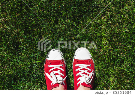 Legs in old red sneakers on green grass. View from above. The concept of youth, spring and freedom Legs in old red sneakers on green grass. View from above. The concept of youth, spring and freedom 85251684