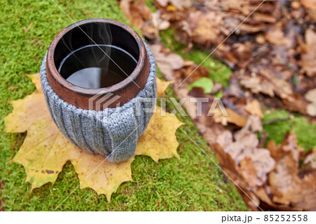 Clay mug in woolen knitted cover. Steam from the mug on the sunny background of autumn forest. Top view. 85252558