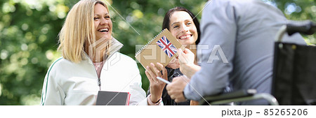 Disabled man in wheelchair giving english textbook to his female friends in park Disabled man in wheelchair giving english textbook to his female friends in park 85265206