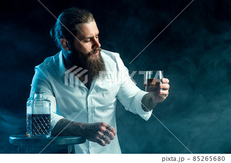 bearded guy holds glass of whiskey, blows smoke on background, dressed in white shirt. Studio shot bearded guy holds glass of whiskey, blows smoke on background, dressed in white shirt. Studio shot 85265680