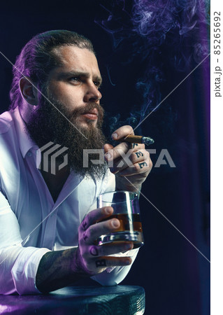 Brutal bearded man smoke a cigar drinks whiskey, blows smoke, dressed in white shirt. Studio shot Brutal bearded man smoke a cigar drinks whiskey, blows smoke, dressed in white shirt. Studio shot 85265692